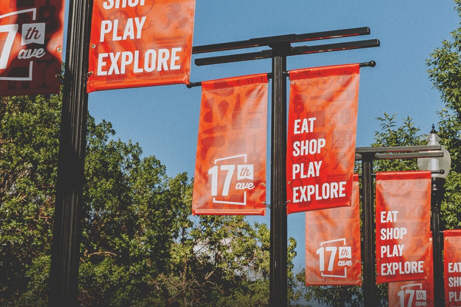 Bright orange street banners reading ‘17th Ave’ and ‘Eat Shop Play Explore’ hang from black lampposts along a tree-lined street under a clear blue sky.