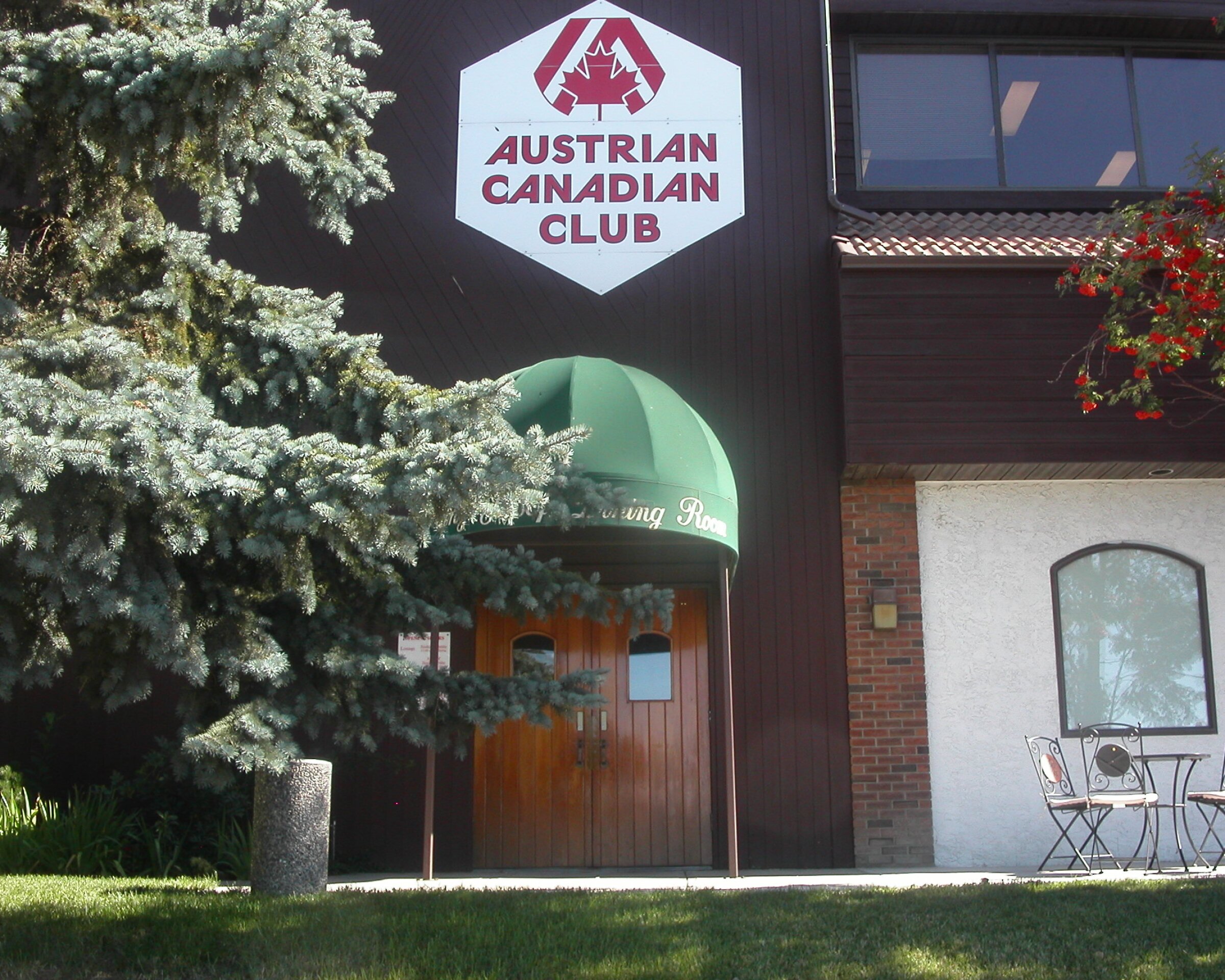 Exterior of the Austrian Canadian Club building, showing a wooden entrance with a green awning, a large sign above the door, and a small patio set on the right side near a lawn and trees.