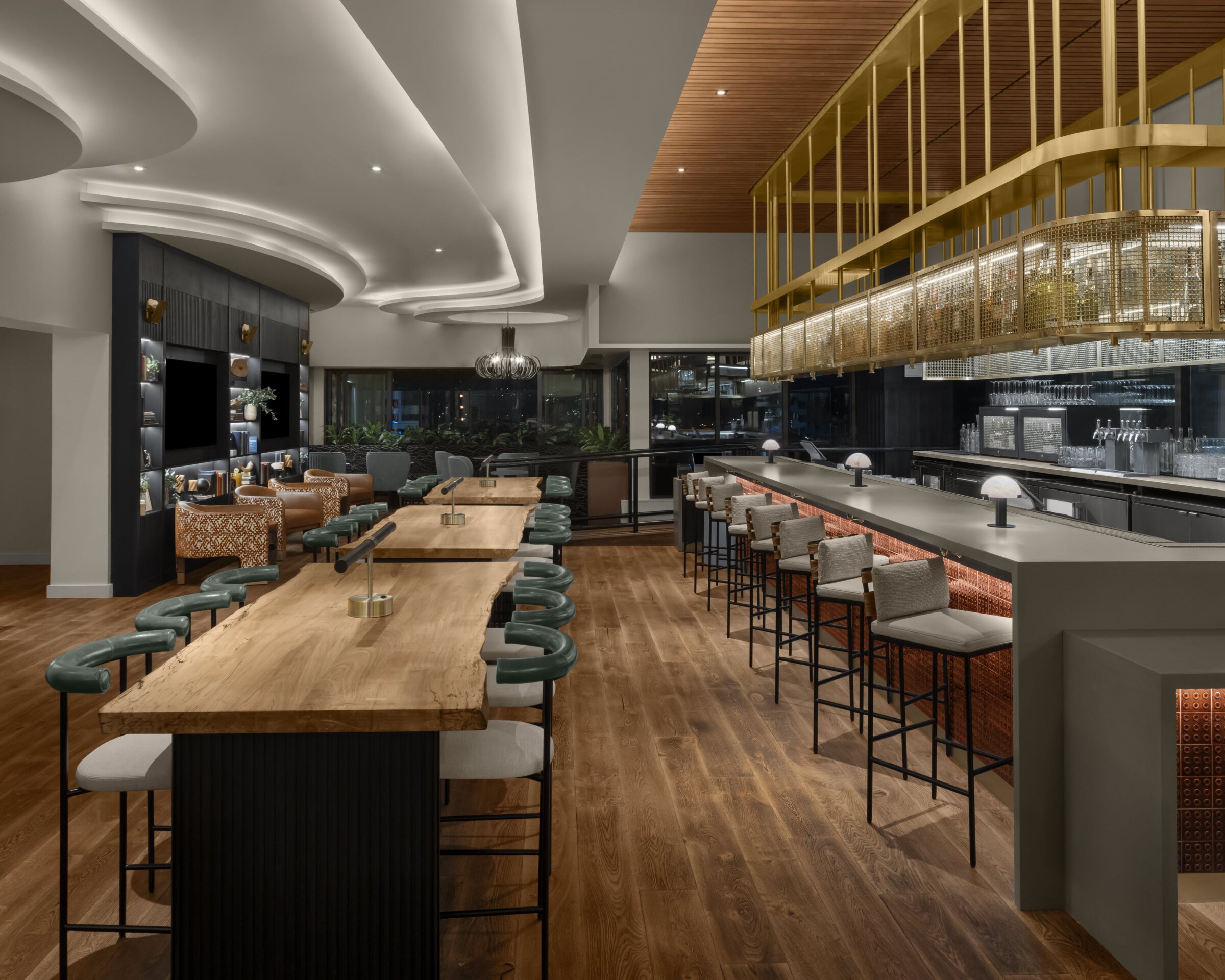 A modern, upscale bar and lounge with wood floors, long communal tables, green upholstered stools, a large bar with high-back chairs, golden overhead shelving, and warm ambient lighting.