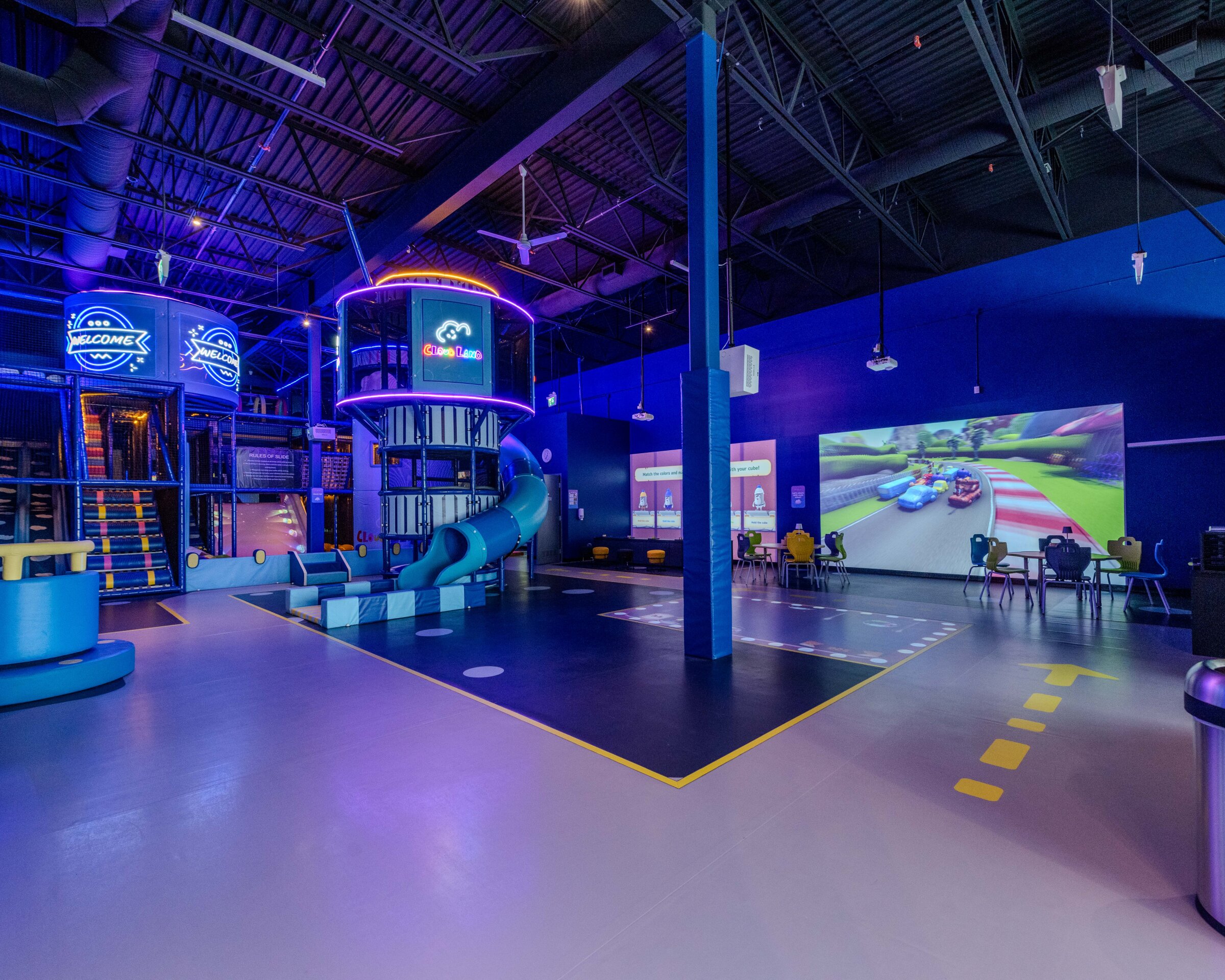 A brightly lit indoor play center with a futuristic, neon-blue theme. There’s a large multi-level climbing structure with glowing signs, a tall central tower with a spiral blue slide, and padded play areas on the floor. On one wall, big projected racing-game screens show colorful cartoon cars. Scattered tables and chairs sit nearby, and the whole space has high industrial ceilings with exposed beams and hanging lights.