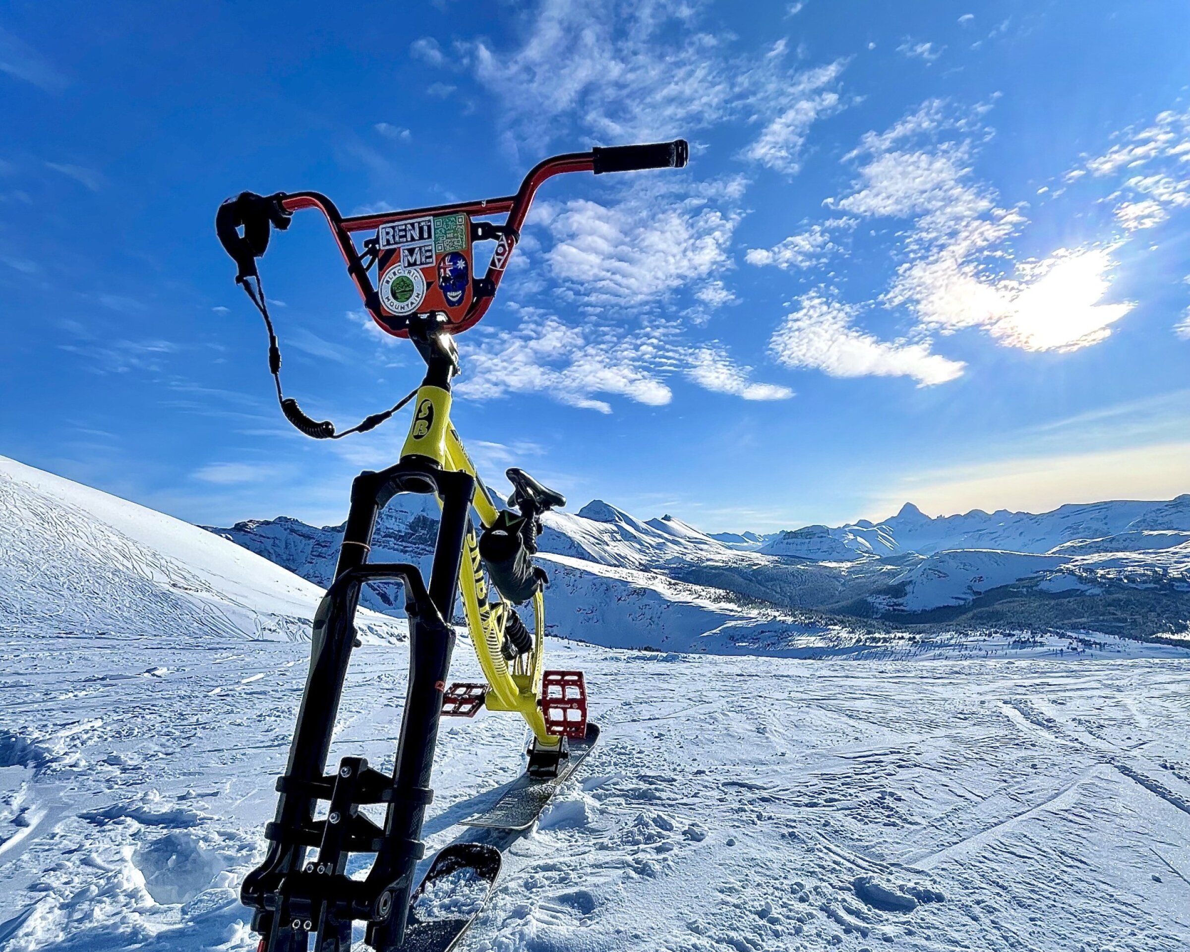 A bright, low-angle shot of a ski bike parked on a snowy mountain slope under a blue sky. The bike’s red handlebars and yellow frame stand out against the white snow, with ski tracks and distant mountain peaks visible in the background. The sun glows near the horizon, lighting up scattered clouds above the ridge line.