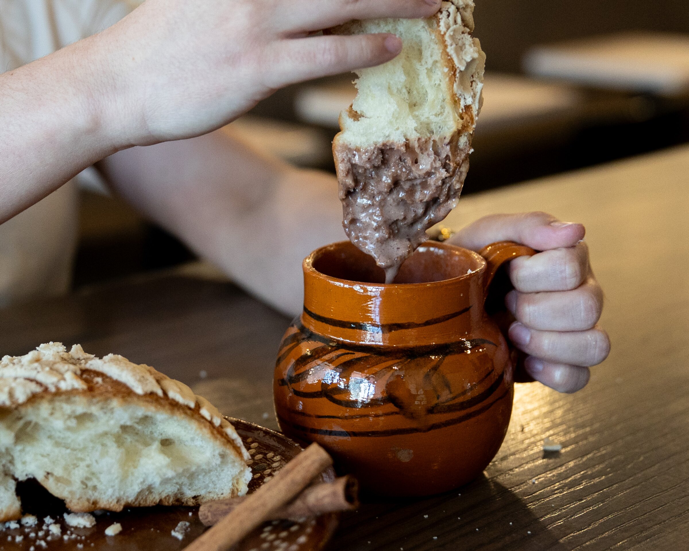 A close-up of someone dipping a thick piece of sweet bread into a clay mug filled with creamy hot chocolate. Another piece of the bread sits on a plate beside two cinnamon sticks on the table.