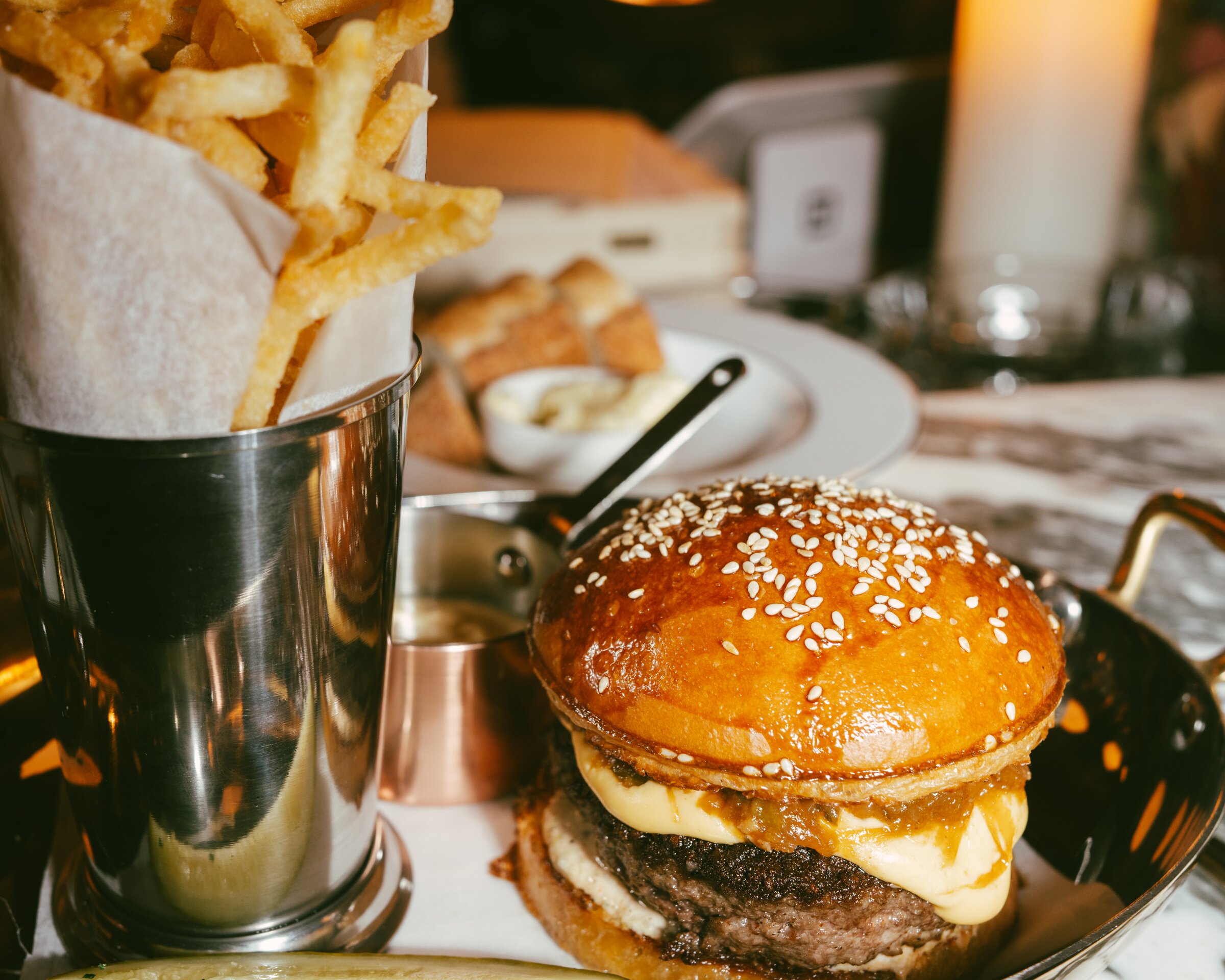 A gourmet burger with melted cheese and caramelized onions sits on a metal tray beside a tall metal cup of fries, a small dipping sauce, and a pickle spear on a marble table in a dimly lit restaurant.