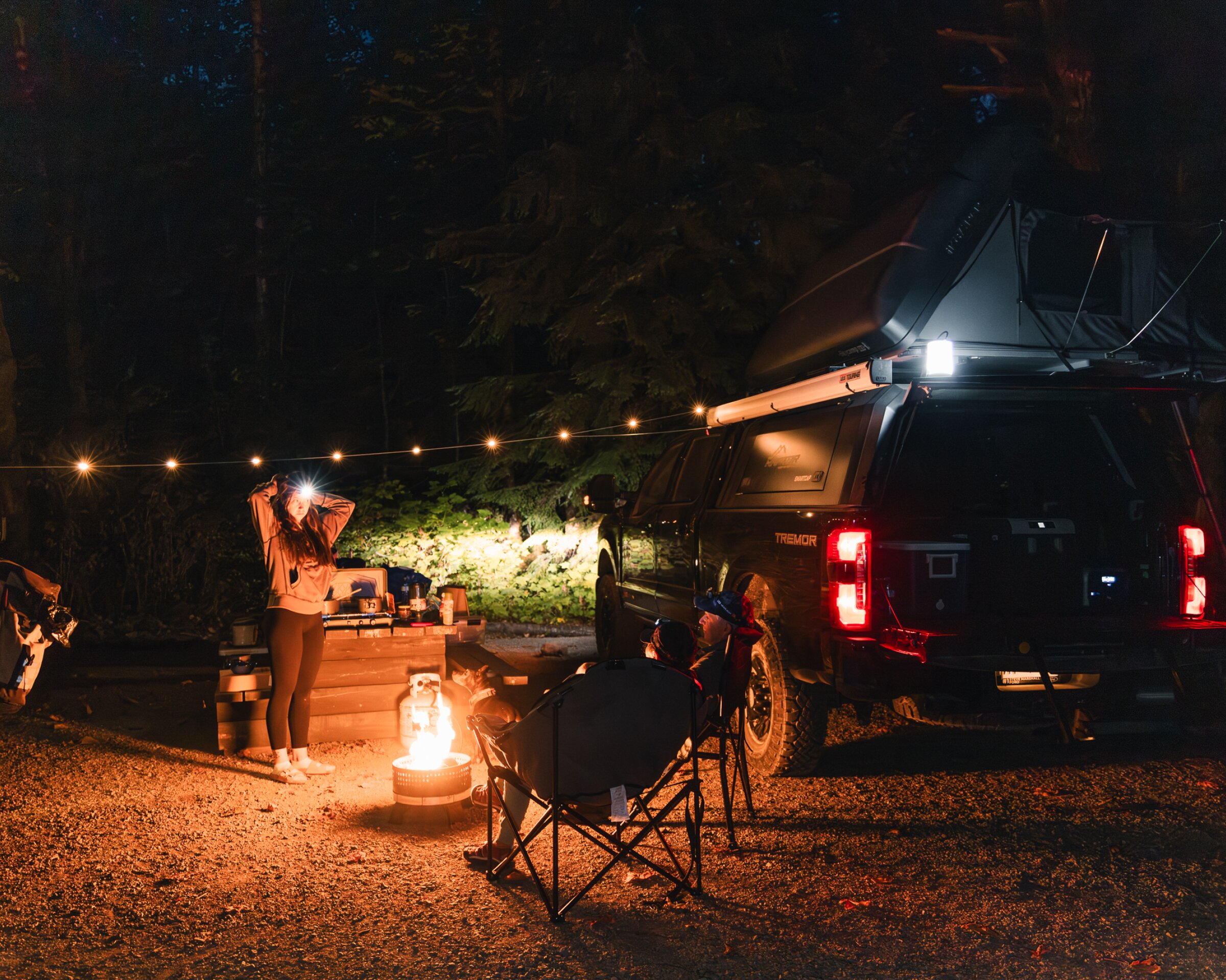 Nighttime campsite in a forest with two vehicles parked beside a picnic table. A rooftop tent is open on a truck, string lights hang between the vehicles, and a small fire burns in a fire pit. One person stands near the table wearing a headlamp, while another sits in a folding chair by the fire. Trees and darkness surround the cozy, well-lit camp setup.