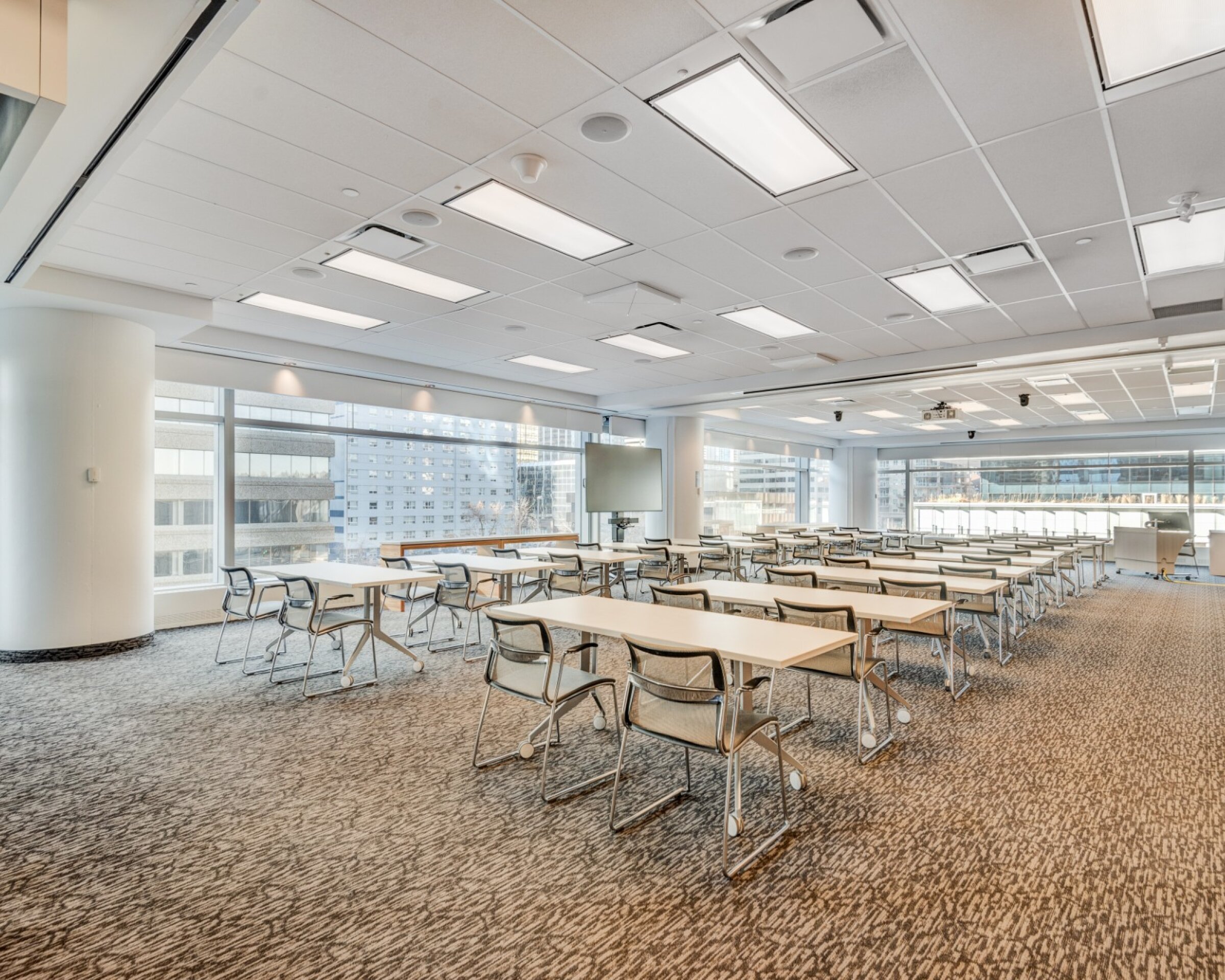 Large, bright conference or classroom-style room with rows of long tables and wheeled chairs, floor-to-ceiling windows along one side, carpeted floors, white columns, and a drop ceiling with recessed lights and mounted screens.