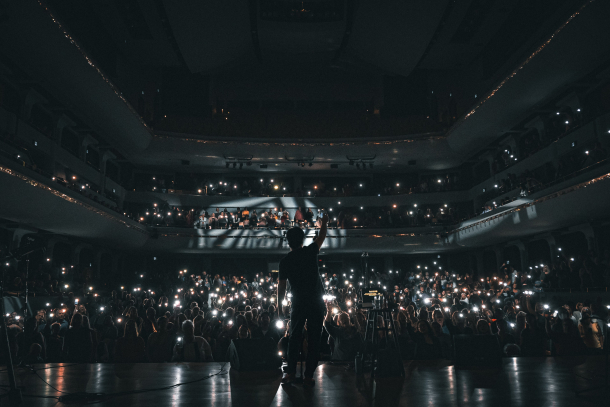 Musician standing on stage facing fans with phone lights shining towards performer