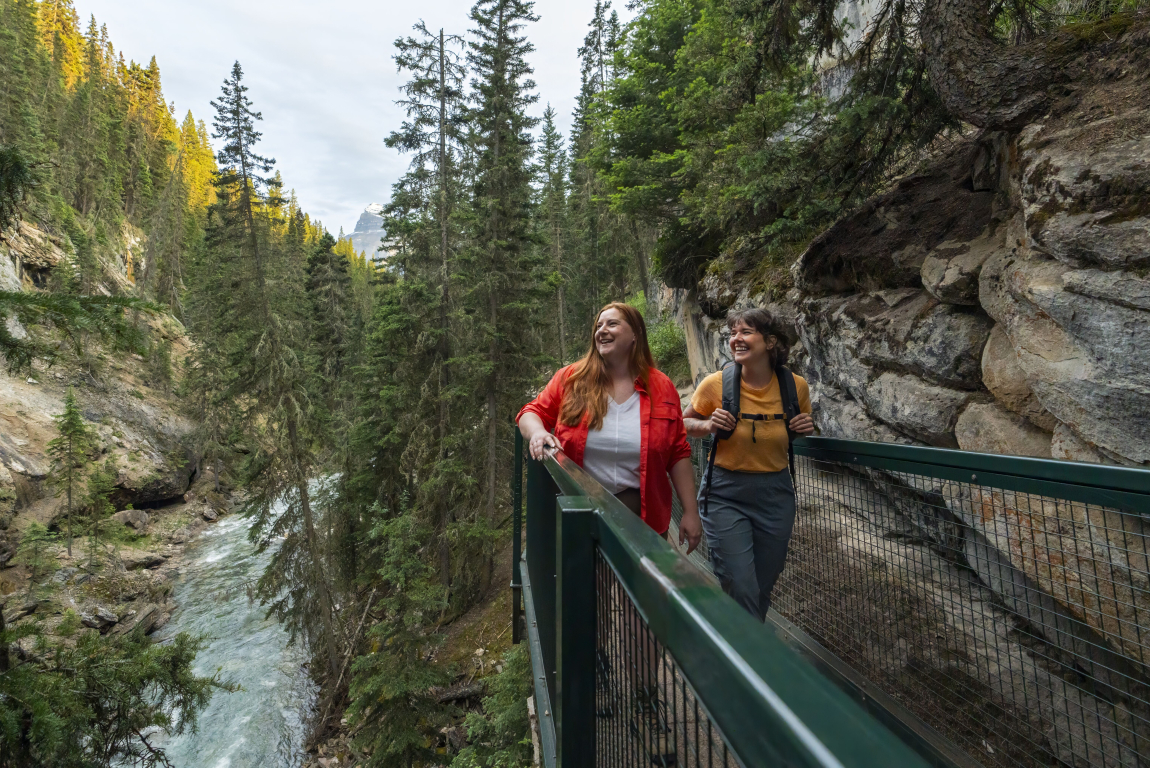 A tight shot of a couple hiking on the boardwalk through Johnston Canyon with the mountain towering in the background; looking out towards the canyon beside them.