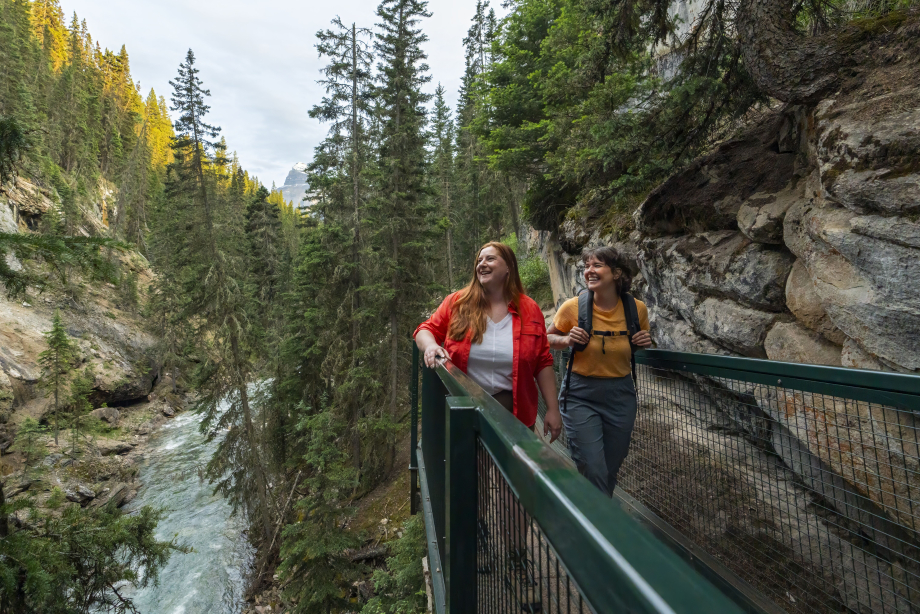 A tight shot of a couple hiking on the boardwalk through Johnston Canyon with the mountain towering in the background; looking out towards the canyon beside them.