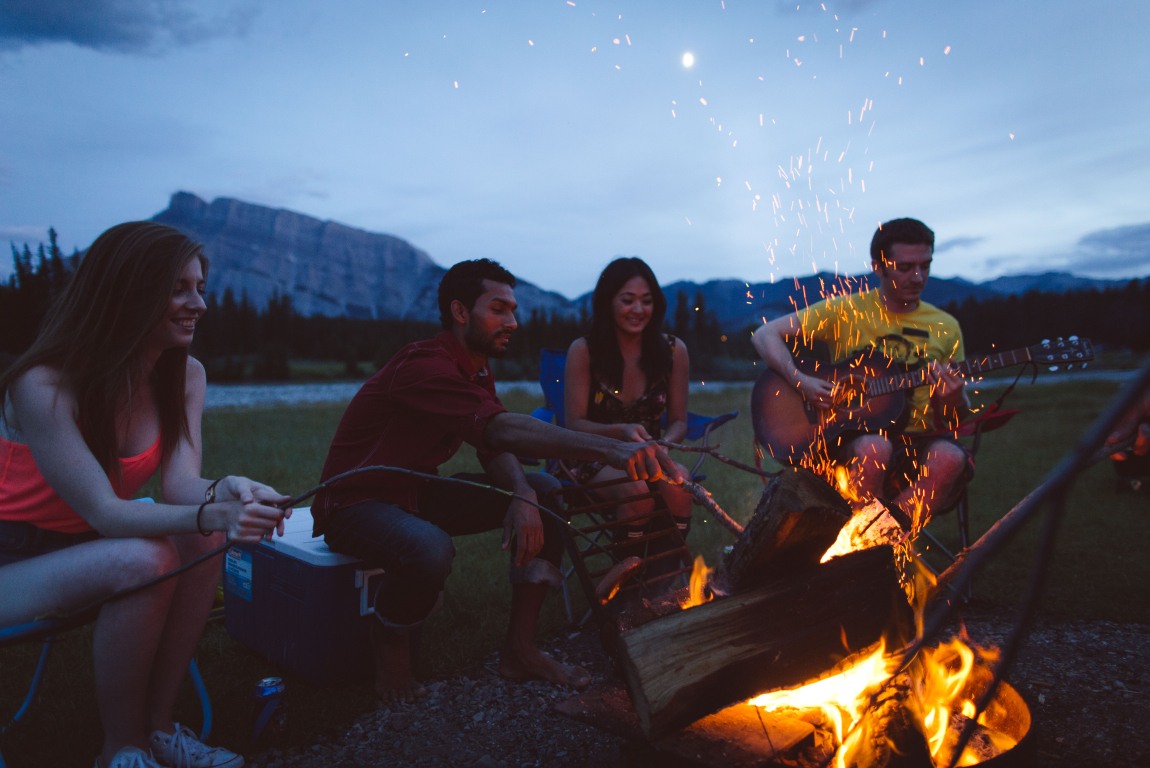 Group of friends playing the guitar around a campfire with mountains and a river in the background