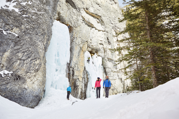 A couple ice walking and hiking through Grotto Canyon while ice climbers scale a rock in the background, near Canmore in Kananaskis Country