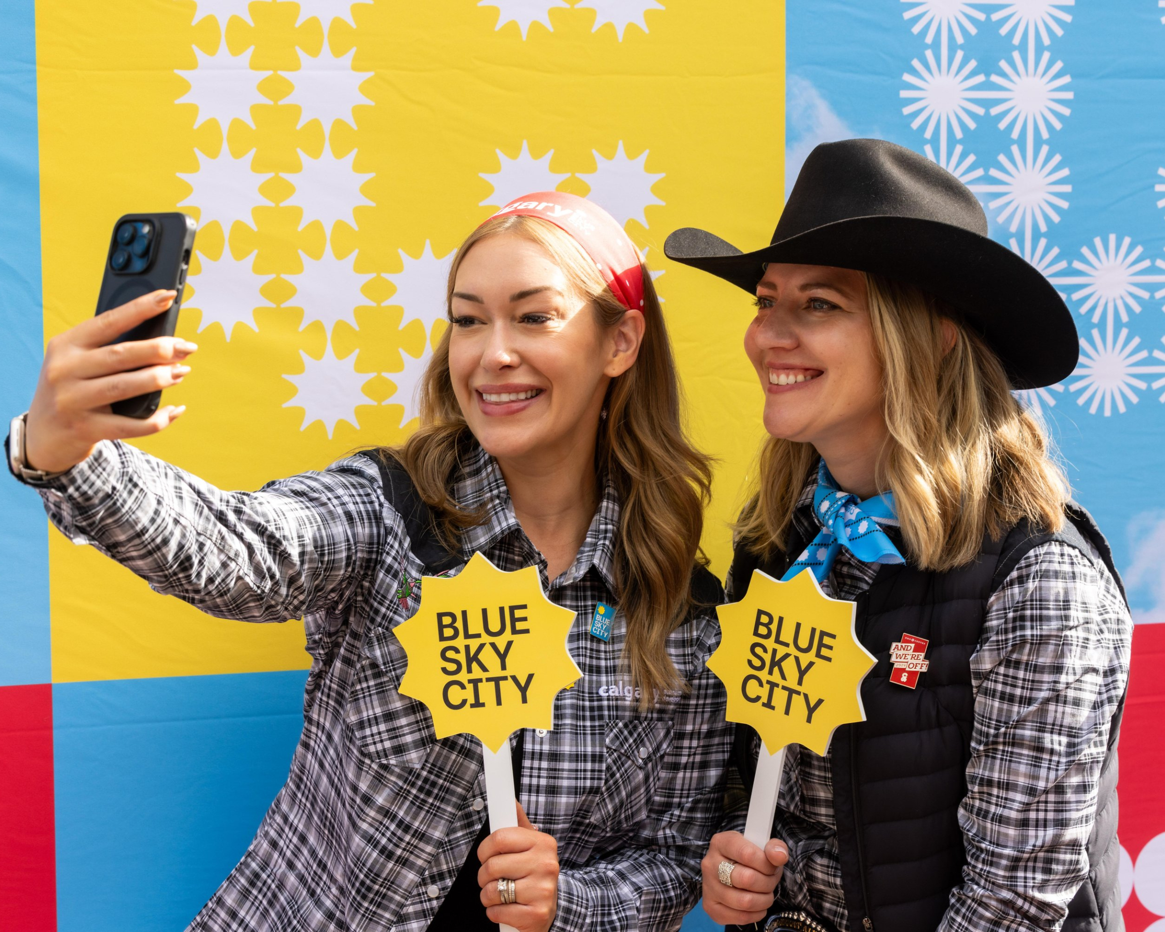 Two women dressed in western attire posing for a selfie with blue sky city paraphernalia