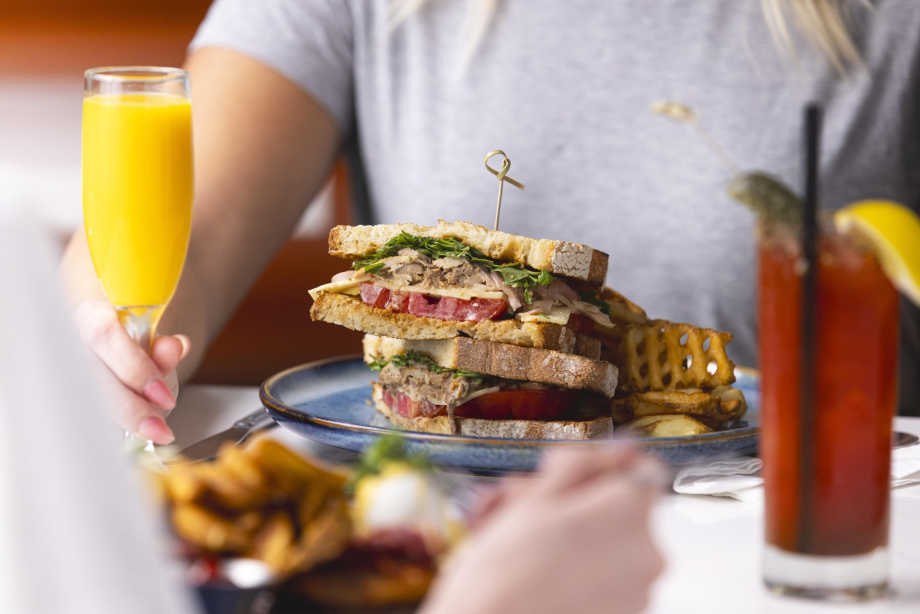 A colourful brunch spread at OEB Breakfast Co. featuring a hearty sandwich, waffle fries, a mimosa and Caesar, served in a bright and lively setting