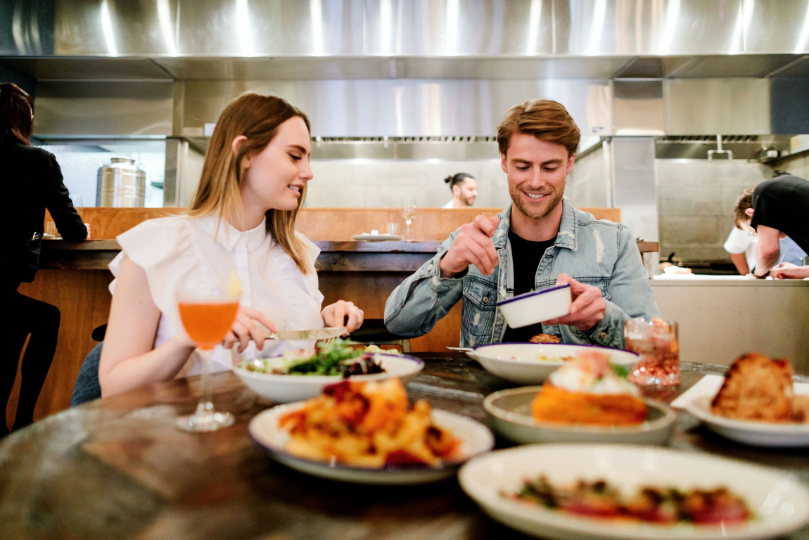 Couple drinking and eating at a restaurant / bar with open kitchen behind them in Calgary