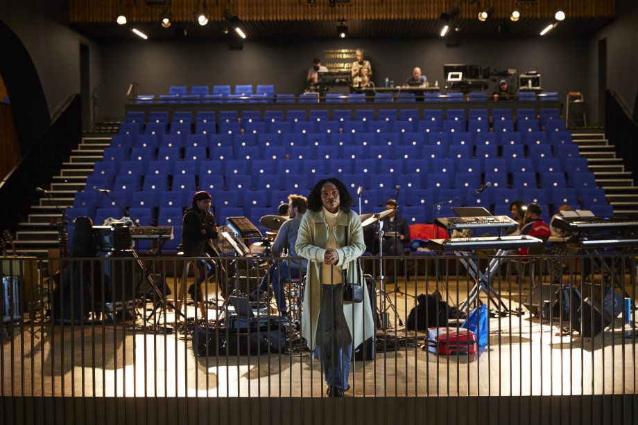 Artist facing the camera with empty seats and band rehearsing behind them