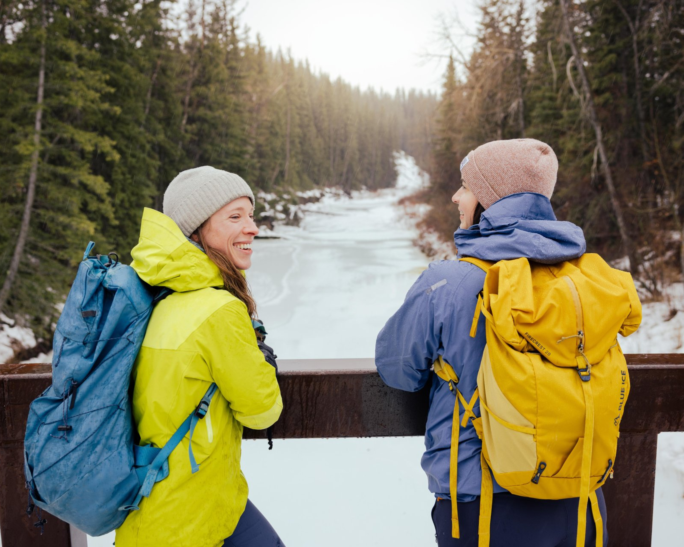 Friends in winter hiking gear overlooking creek from a bridge.