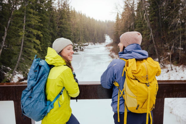 Friends in winter hiking gear overlooking creek from a bridge.