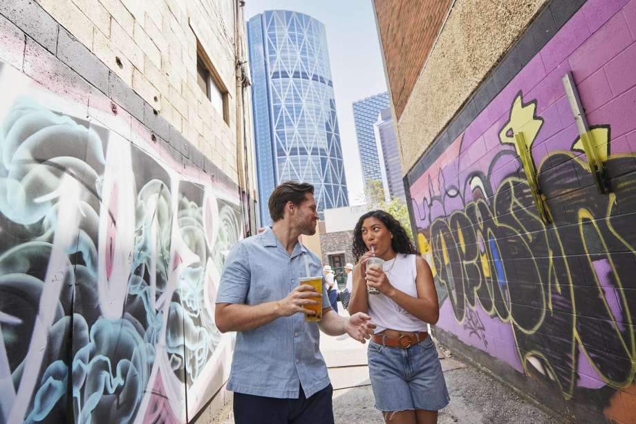 couple sipping bubble tea and walking through mural alley in Calgary's Chinatown, with the Bow tower behind them peeking between two buildings