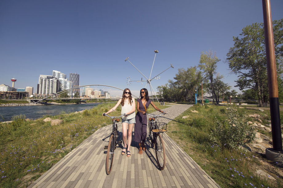 Two women walking their bikes along the Riverwalk on St. Patrick's Island with Calgary Tower in background