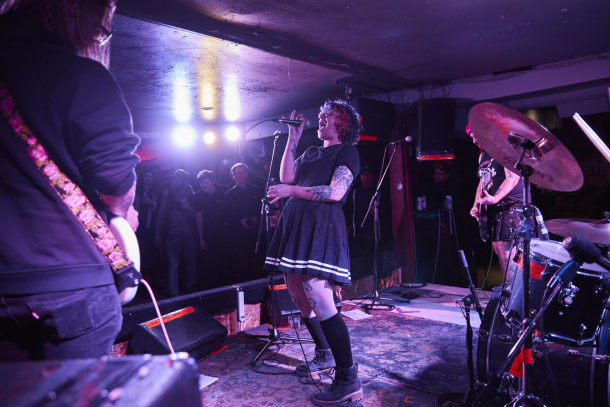 Woman singing into mic on stage with guitarists and fans in the background