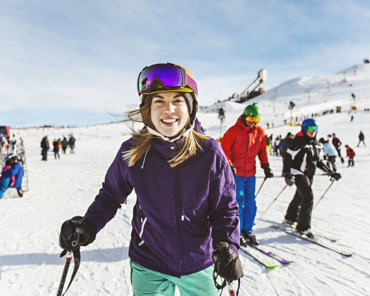 A women standing at the base of the hill, smiling into the camera, with skiers and boarders in the background, before skiing at WinSport in Calgary